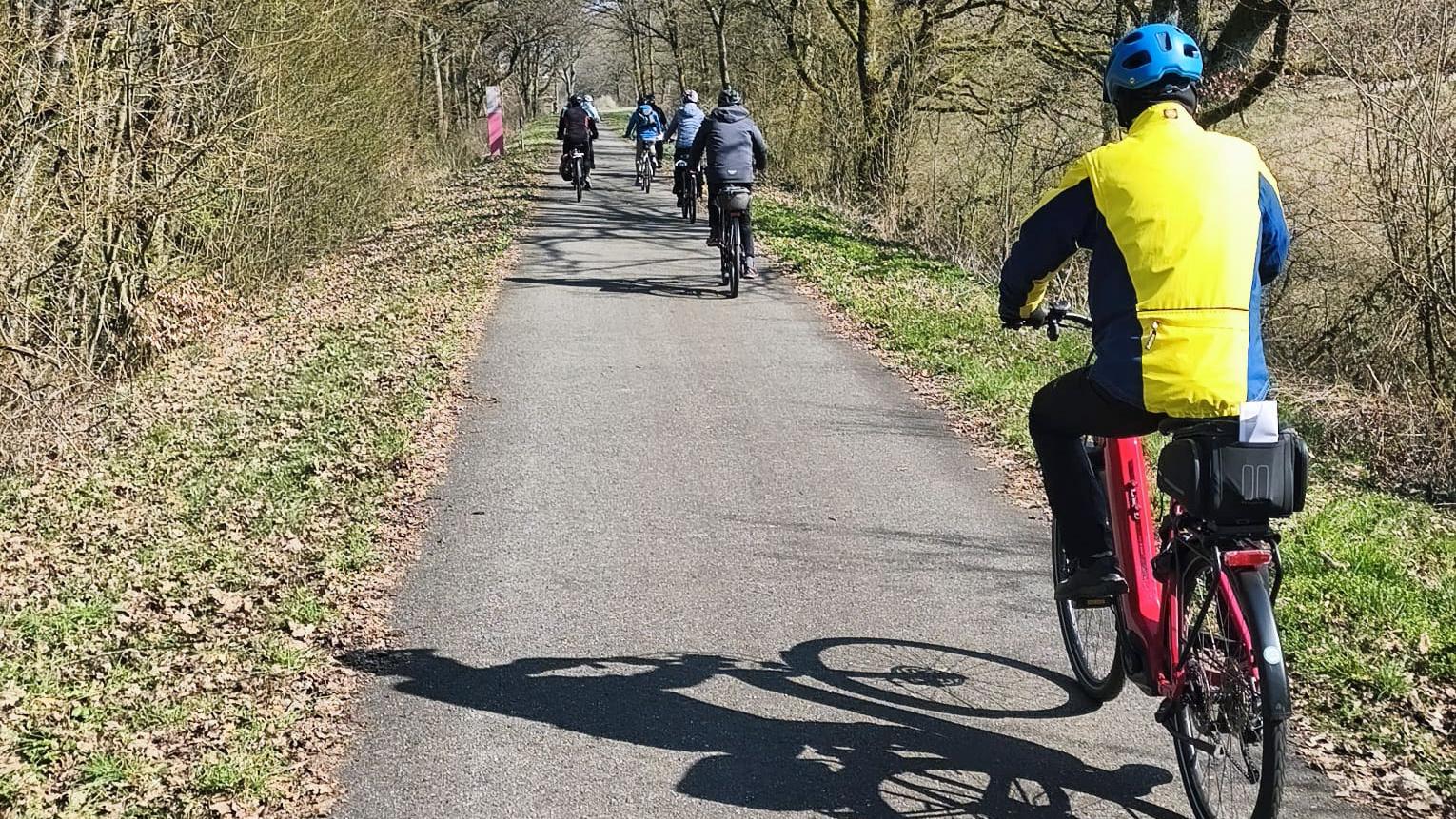 Mehrere Personen fahren mit Fahrrädern auf einem asphaltierten Weg durch eine baumreiche Landschaft. Der Weg ist von kahlen, frühsaisonalen Bäumen und niedrigem Gebüsch gesäumt. Im Vordergrund fährt eine Person auf einem roten E‑Bike, getragen wird ein blau‑gelber Fahrradhelm und eine passende Jacke. Weiter vorn auf dem Weg radelt eine Gruppe in lockerer Formation. Die Sonne wirft deutliche Schatten der Fahrräder auf den Boden. Rechts und links des Weges liegen trockene Blätter und Grasflächen, die Umgebung wirkt ruhig und ländlich.