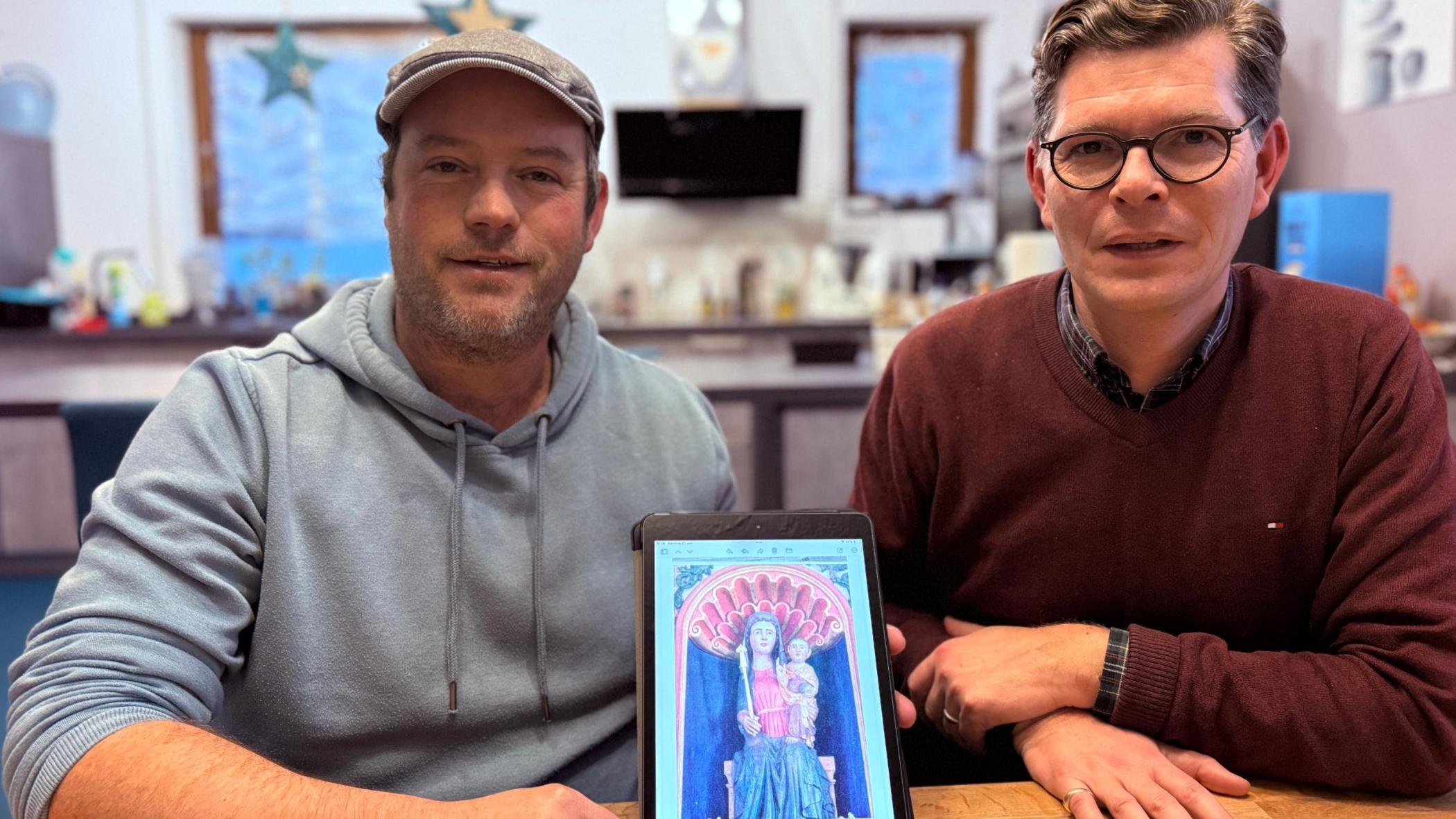 Zwei Männer an einem Tisch zeigen ein Tablet mit dem Foto einer Madonnen-Figur.