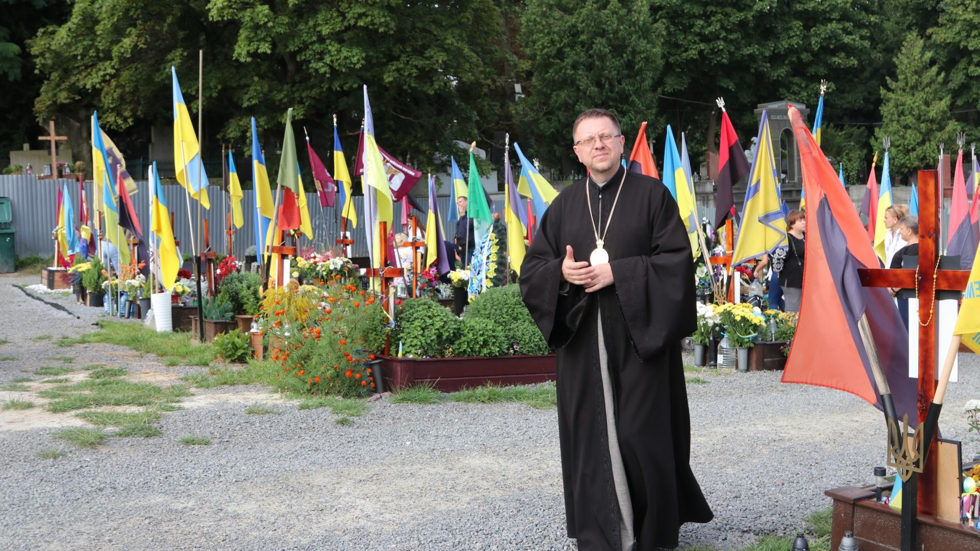 Mann mit Soutane in schwarz steht zwischen Fahnen auf einem Friedhof