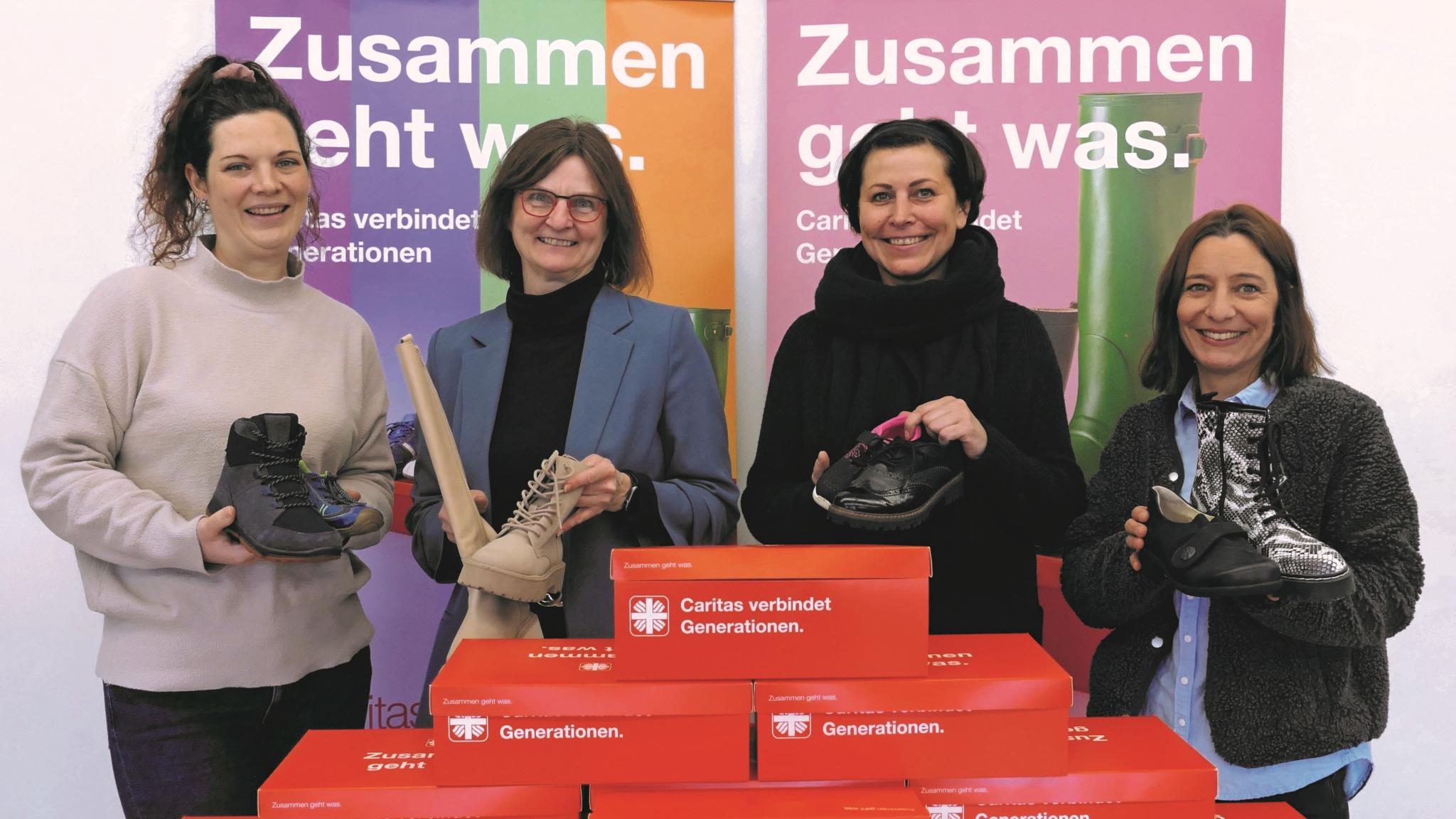 Vier Frauen lächeln in die Kamera und halten je einen Schuh hoch, vor ihnen stehen rote Schuhkartons.