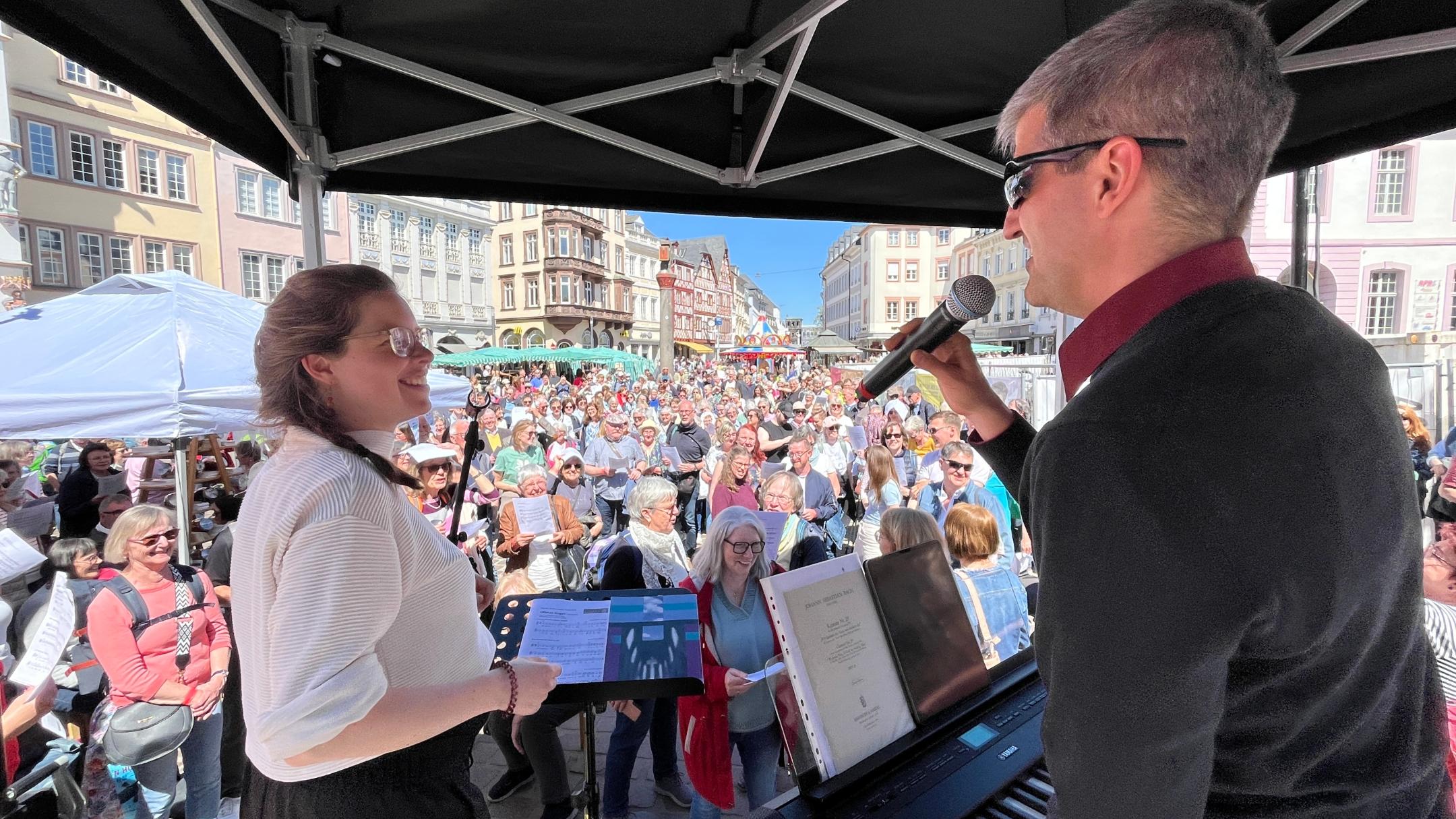 Eine Frau und ein Mann haben Mikrofone in der Hand, der Mann spielt Keyboard und sie stehen auf einer Bühne vor der sehr viele Leute auf einem Platz stehen.