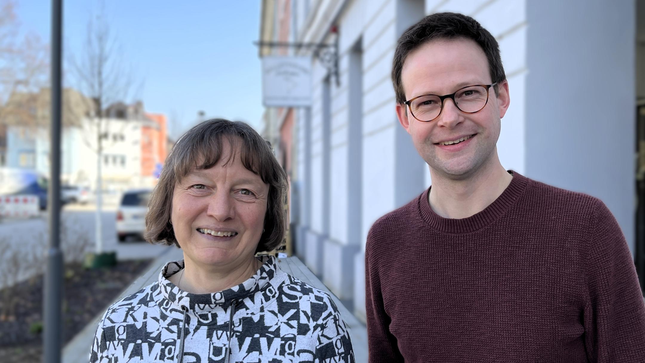 Eine Frau und ein junger Mann lachen in die Kamera und stehen in einer Einkaufsstraße.