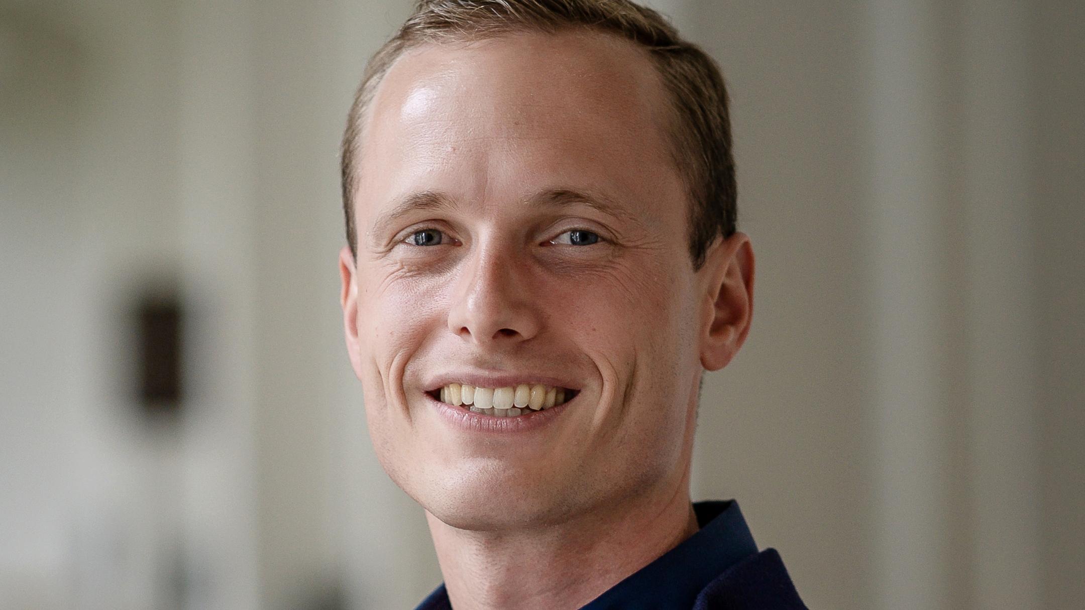 Mann in blauem Pullover steht in einem hellen Raum und lächelt in die Kamera