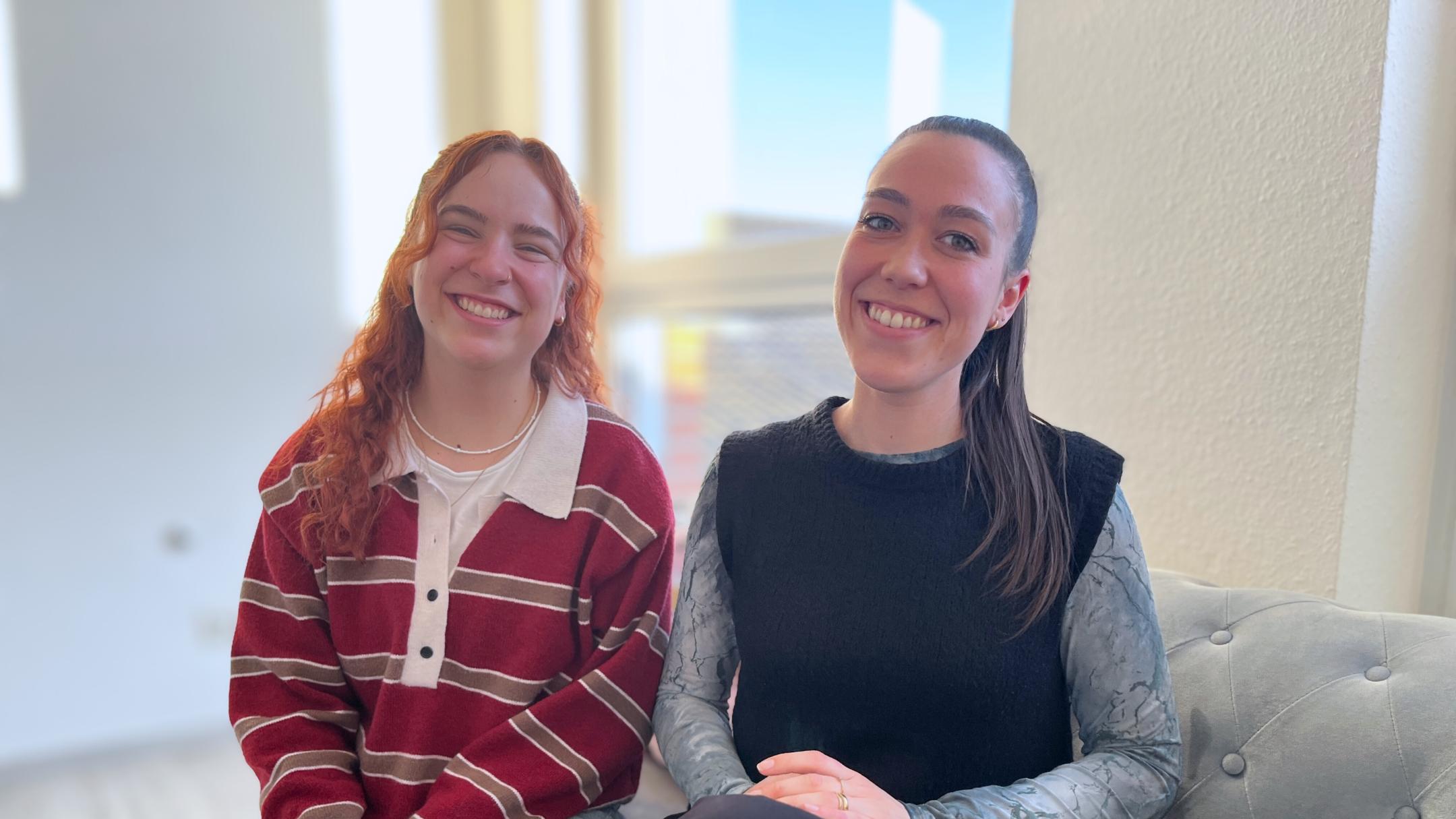 Zwei junge Frauen sitzen nebeneinander auf einem Sofa und lachen in die Kamera.