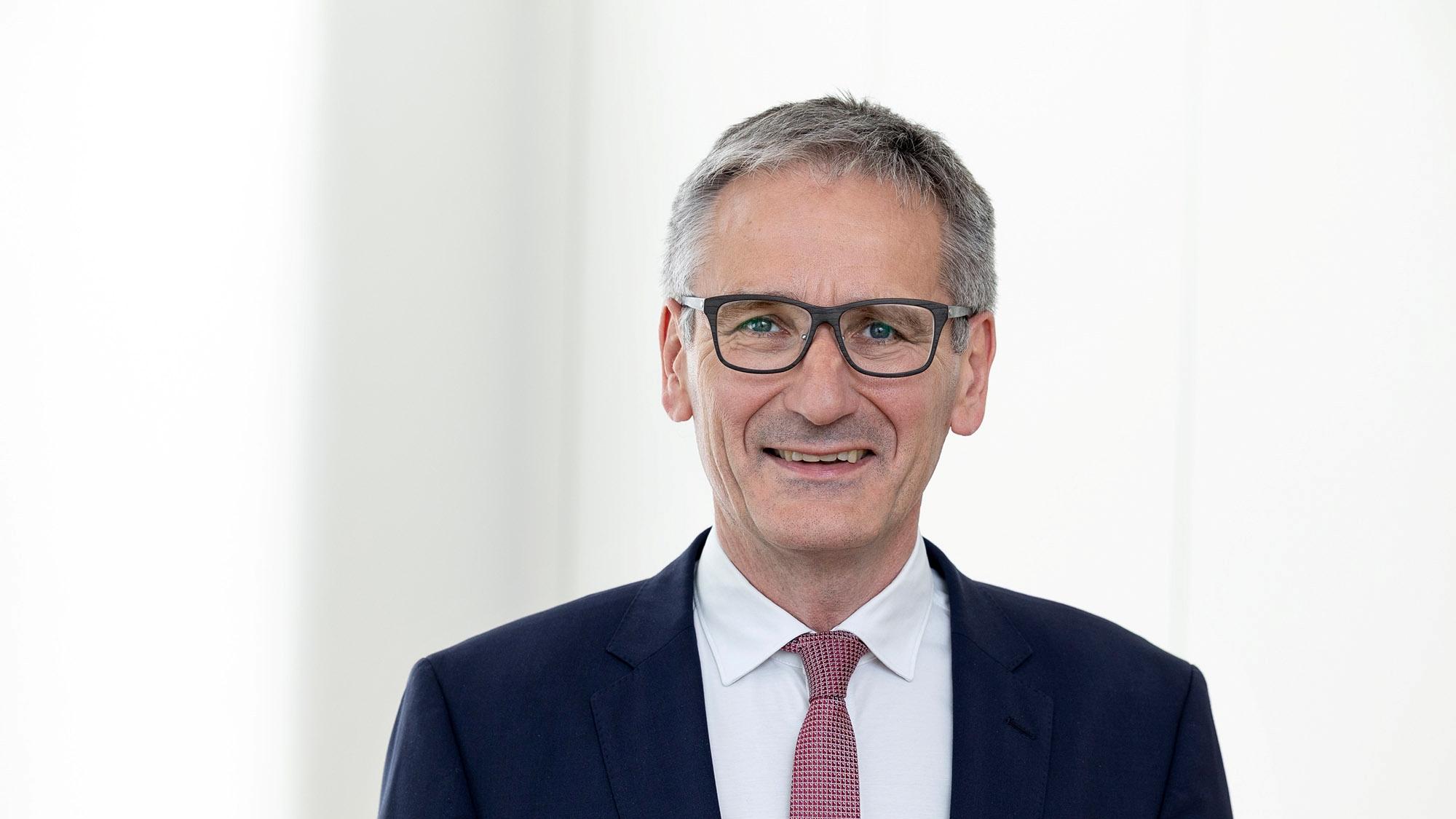 Mann in dunklem Anzug vor hellem Hintergrund blickt geradeaus in die Kamera.