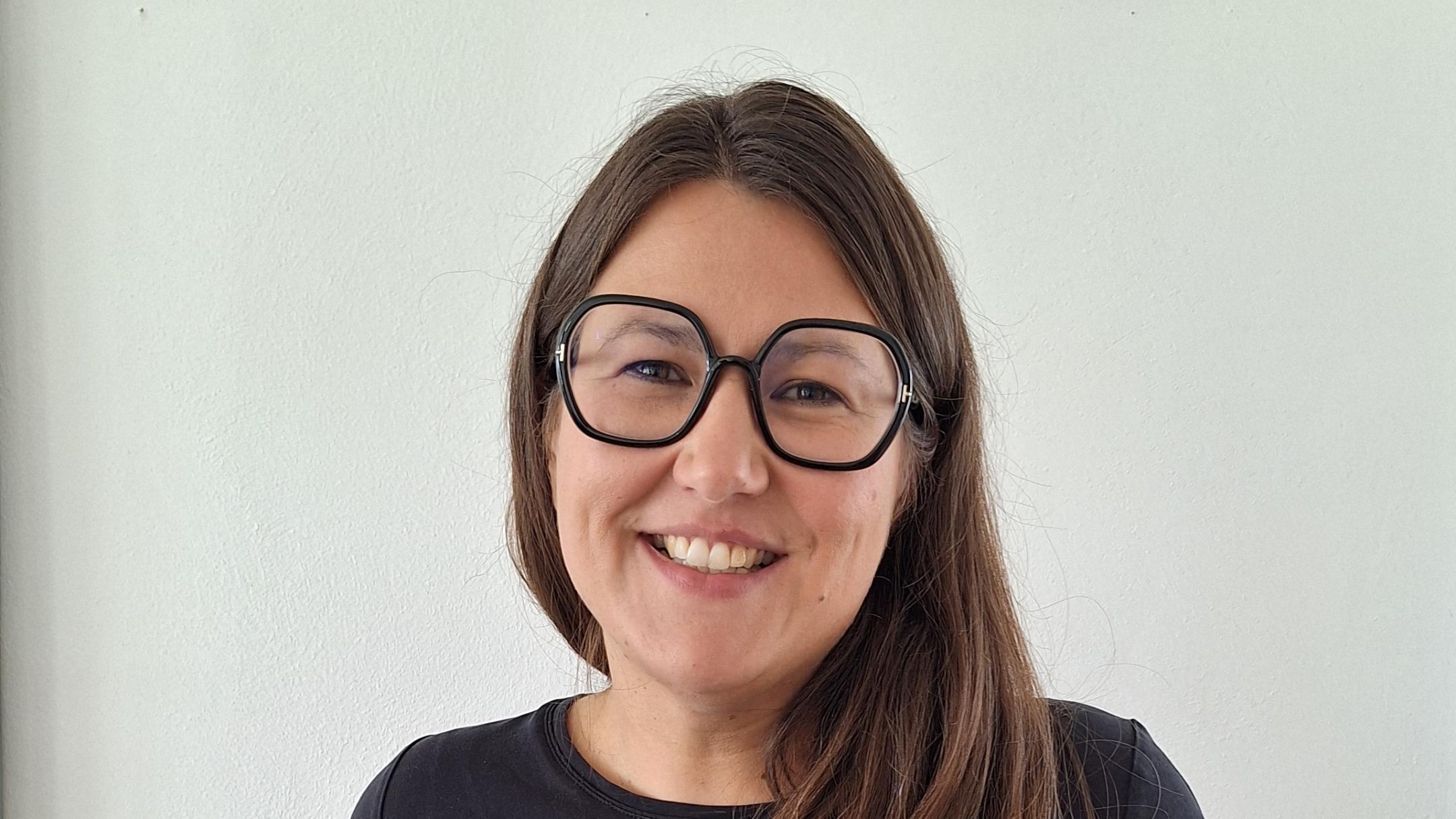 Junge Frau mit langen Haaren, runder Brille und T Shirt steht vor weißer Wand.